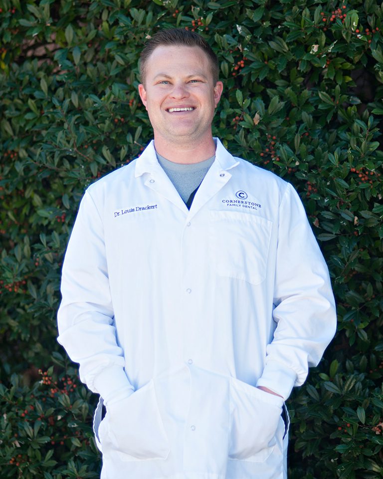 A man in white lab coat standing next to green bushes.